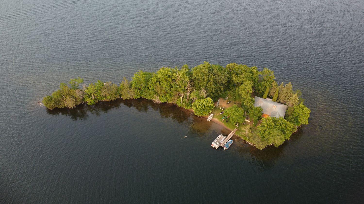 A teardrop shaped island with cabin, thick forest, and dock sit in dark grey waters of Fish Trap Lake